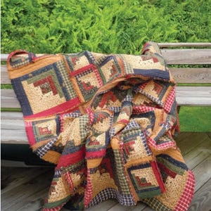 May include: A patchwork quilt with a rustic, country aesthetic. The quilt features a log cabin design with squares in shades of tan, red, green, and blue. The quilt is draped over a wooden bench, with a backdrop of green foliage.