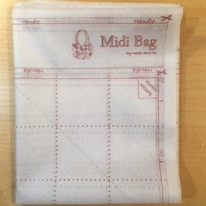 May include: A white fabric pattern for a "Midi Bag" with red printed lines and text. The design includes dotted lines, a basket illustration, and the word "Handle" repeated. The pattern is folded and lies on a light brown surface.
