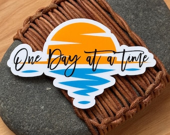 May include: A sticker with a sunset and ocean scene and the text "One Day at a Time".