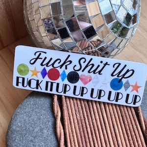 Puede incluir: Una pegatina blanca con el texto "Fuck Shit Up" en escritura negra. Debajo, las palabras "Fuck It Up Up Up Up Up" en negro, con formas geométricas coloridas. Una bola de discoteca en el fondo.