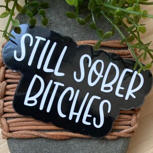May include: Black and white sticker with the text "Still Sober Bitches" in a white cursive font.