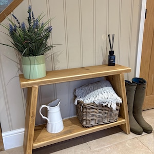 May include: A light oak entryway bench with a lower shelf. The top shelf holds a green potted plant with purple flowers and a black diffuser. The lower shelf displays a white jug, a wicker basket with a grey blanket, and olive green wellington boots.