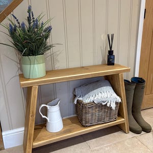 May include: A wooden console table with a lower shelf, featuring a light green pot with artificial lavender, a black reed diffuser, a white ceramic pitcher, a woven wicker basket containing a grey and white chevron throw, and two tall olive boots.