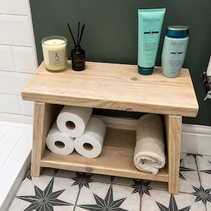 May include: A light-toned wooden bathroom bench with a top shelf and a lower storage shelf. The top holds a yellow candle, a reed diffuser, and two Kérastase Resistance hair care bottles. The lower shelf holds three toilet paper rolls and a towel.