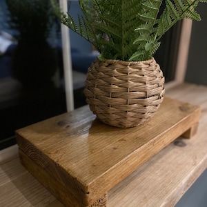 May include: A wooden plant stand with a natural brown finish and a woven wicker planter with a green fern plant. The stand has a rectangular top and four legs.