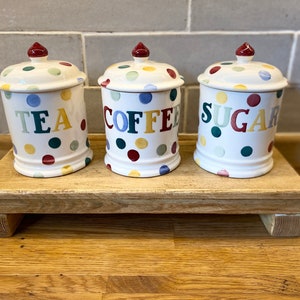 May include: Three white ceramic canisters with lids, each featuring colourful polka dots and the words TEA, COFFEE, and SUGAR printed in multi-coloured letters. Each lid is topped with a small, dark red knob. The canisters are arranged in a row.