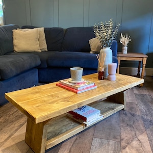 May include: A light-wood coffee table with a lower shelf, holding books. The table is rectangular and has a natural finish. A cup, books, and decorative items are on top. A blue sofa and wall are in the background. The table is approximately 120 cm long.
