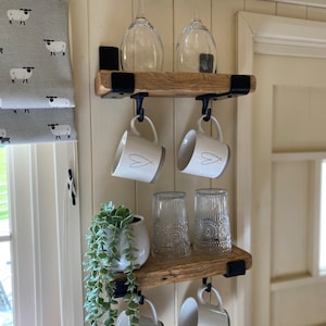 May include: Two stacked wooden shelves with black metal brackets and hooks. The top shelf holds two clear wine glasses. The bottom shelf holds a small potted plant and two clear textured glasses. Four white mugs with heart designs hang from hooks.