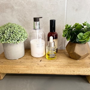 May include: A rectangular wooden riser with four short feet, holding a grey pot with a green faux plant, a clear Molton Brown hand lotion bottle, a small Clarins oil bottle with a dropper, a dark brown spray bottle, and a faceted gold pot with a plant.