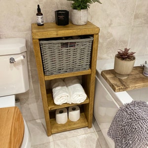 May include: A wooden bathroom shelf with three shelves. The top shelf has a grey wicker basket, a black candle, and a small bottle. The middle shelf has two white towels. The bottom shelf has two rolls of toilet paper. The shelf is in a bathroom with a white toilet and a white bath.