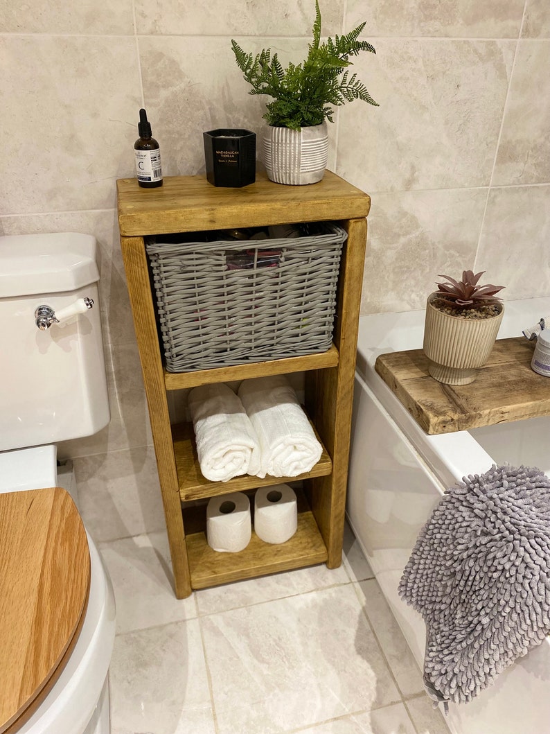 May include: A wooden bathroom shelf with three shelves. The top shelf has a gray wicker basket, a black candle, and a small bottle. The middle shelf has two white towels. The bottom shelf has two rolls of toilet paper. The shelf is in a bathroom with a white toilet and a white bathtub.
