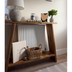 May include: A wooden console table with an A-frame design, featuring a top surface and a lower shelf. The table is made of medium-toned wood with visible grain. It has angled side supports that connect the top and bottom shelves, creating a sturdy structure.