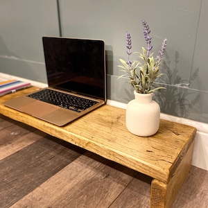 May include: A rectangular wooden monitor stand with a natural grain finish, featuring a laptop computer with a black screen and a small white ceramic vase containing artificial purple lavender sprigs placed on its surface.