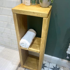 May include: A wooden shelf unit with two shelves, a white towel, a bottle of hair product labeled "Olaplex No. 3 Hair Perfector", and a bottle of scented room spray labeled "Eucalyptus & Cedarwood" on the top shelf. The shelf unit is in a bathroom with white tile walls and a green wall behind it.