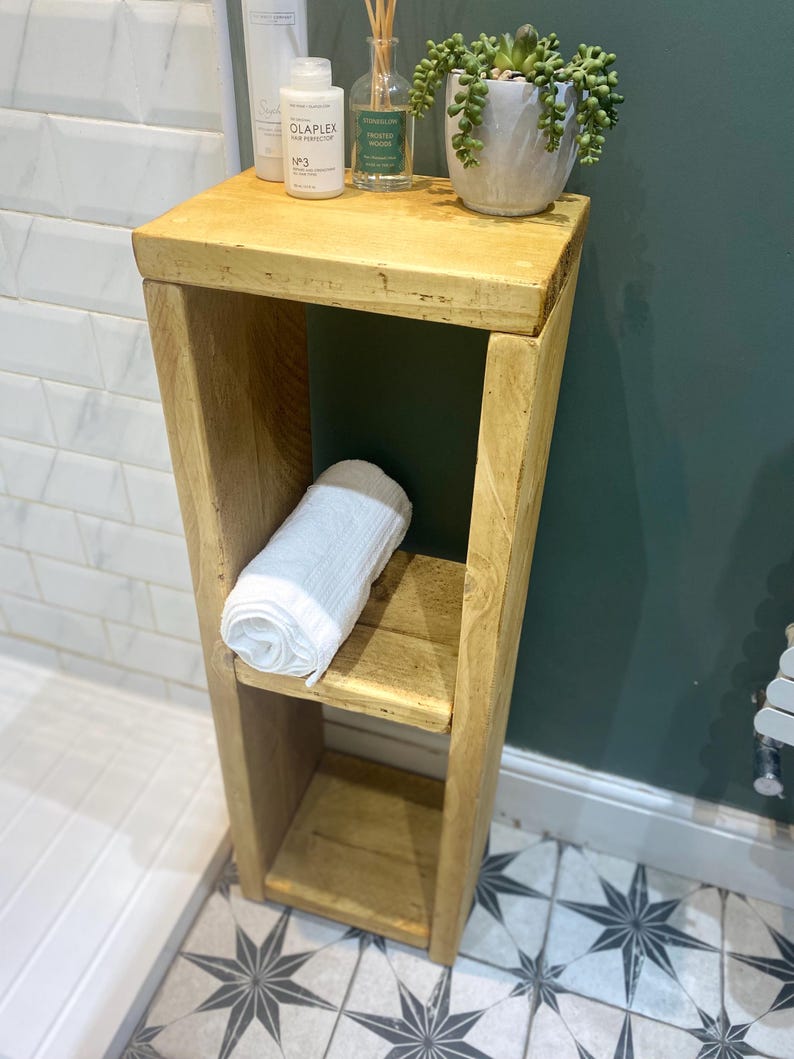 May include: A wooden shelf unit with two shelves, a white towel, a bottle of hair product labeled "Olaplex No. 3 Hair Perfector", and a bottle of scented room spray labeled "Eucalyptus & Cedarwood" on the top shelf. The shelf unit is in a bathroom with white tile walls and a green wall behind it.