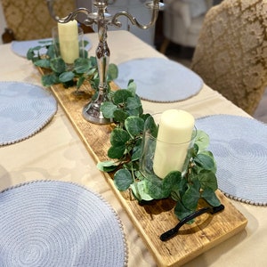 May include: A wooden serving tray with a black handle, decorated with greenery and candles. The tray is set on a table with blue placemats.