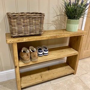 May include: A rectangular wooden shoe rack with two shelves featuring a natural light brown finish. A woven wicker basket sits on the top shelf next to a light green pot containing artificial lavender. A pair of tan boots and trainers are on the shelf.