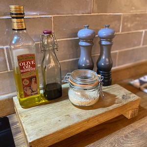 May include: A wooden kitchen shelf displays a bottle of olive oil, a small glass bottle, a jar of spices, and two grey pepper grinders. The olive oil bottle has a gold cap and a label. The shelf is made of light-coloured wood.