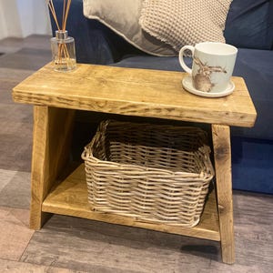 May include: A small, wooden side table with a lower shelf and a wicker basket. A mug with a rabbit illustration sits on a saucer. A glass diffuser with reeds is also on the table.