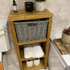 May include: A wooden bathroom storage shelf with three shelves. The top shelf has a gray wicker basket, a black candle, and a small bottle of liquid. The middle shelf has two white towels rolled up. The bottom shelf has two rolls of toilet paper. The shelf is in a bathroom with white tile floors and a white bathtub.