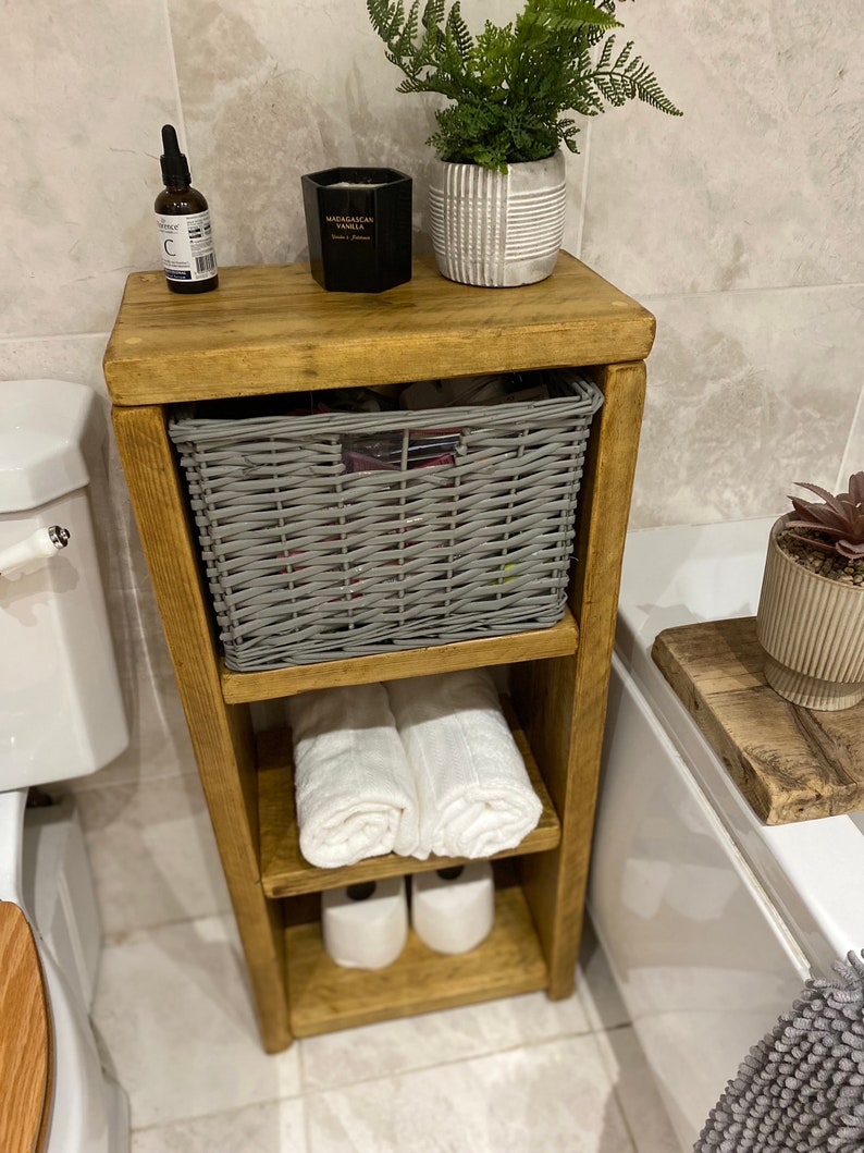 May include: A wooden bathroom storage shelf with three shelves. The top shelf has a gray wicker basket, a black candle, and a small bottle of liquid. The middle shelf has two white towels rolled up. The bottom shelf has two rolls of toilet paper. The shelf is in a bathroom with white tile floors and a white bathtub.