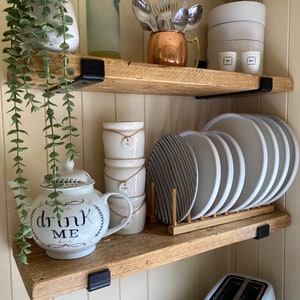 May include: Two rustic wooden shelves with black metal brackets holding kitchen items. Top shelf: a plant, glass bottle, copper mug with silverware, and white bowls. Bottom shelf: a teapot with 'drink me' text, stacked canisters, and plates in a rack.