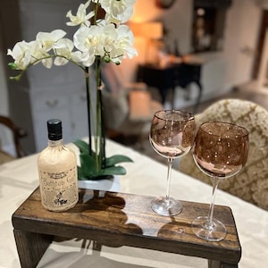 May include: A dark brown wooden riser stand holding a bottle of Bathtub Gin with a cream-coloured paper label and two stemmed wine glasses with tinted amber bowls. The bottle features black text and a botanical illustration on the front label.