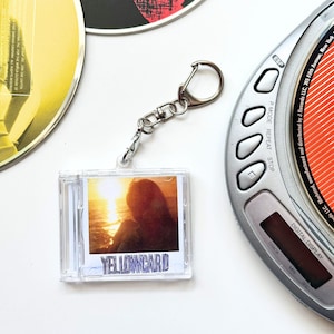 May include: A clear plastic miniature CD case keyring featuring the Yellowcard album cover art with a sunset over the ocean. The keyring includes a silver-toned metal lobster clasp and jump ring, positioned next to a portable CD player.