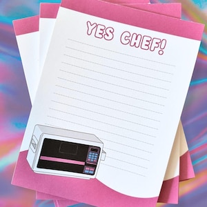 May include: A stack of notepads with a pink border and the text "YES CHEF!" in pink. Each notepad has lined paper and a graphic of a microwave oven. The background has a holographic effect.