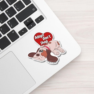 May include: A sticker showing a cartoon dog with brown and white fur, wearing a red collar, and lying down. Above the dog is a red heart with the words "Adopt Don't Shop". The sticker is on a white laptop.