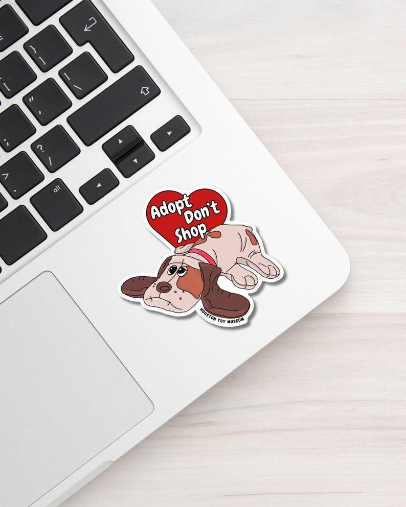 May include: A sticker showing a cartoon dog with brown and white fur, wearing a red collar, and lying down. Above the dog is a red heart with the words "Adopt Don't Shop". The sticker is on a white laptop.