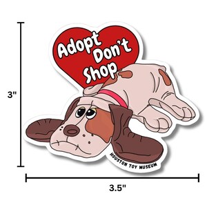 May include: A sticker featuring a cartoon dog with brown and tan fur, wearing a red collar. The dog is lying down with a sad expression. Above the dog is a red heart with the words "Adopt Don't Shop". The sticker is 7.6 cm tall and 8.9 cm wide.