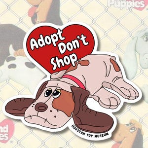 May include: A sticker depicting a cartoon dog with brown and white fur, wearing a red collar, and a red heart with the words "Adopt Don't Shop." The sticker is from the Houston Toy Museum and has a white border.