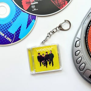 May include: A square keychain featuring a miniature replica of the Weezer self-titled album cover, encased in a clear plastic jewel case. The artwork displays the band members against a bright yellow background with the word Weezer in white text.