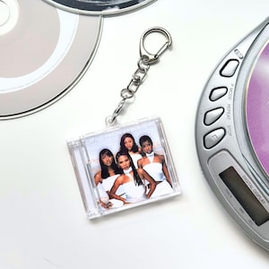 May include: A square clear acrylic keychain featuring a miniature replica of a CD jewel case. The insert displays a photograph of four individuals wearing white tops. The keychain includes a silver-toned metal lobster clasp and chain.