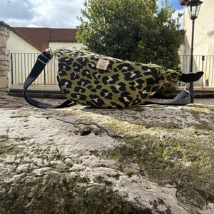 Op de afbeelding: Een heuptasje met luipaardprint in olijfgroen en zwart, met een zwarte verstelbare riem. De tas heeft een klein label. De tas ligt op een stenen oppervlak.