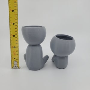 Puede incluir: Dos macetas de cer&aacute;mica gris con forma de figuras humanas. La maceta de la izquierda est&aacute; sentada erguida con los brazos cruzados. La maceta de la derecha est&aacute; sentada con las piernas cruzadas.