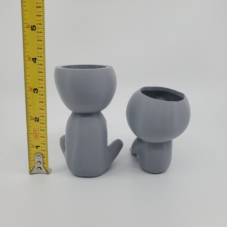 Puede incluir: Dos macetas de cer&aacute;mica gris con forma de figuras humanas. La maceta de la izquierda est&aacute; sentada erguida con los brazos cruzados. La maceta de la derecha est&aacute; sentada con las piernas cruzadas.