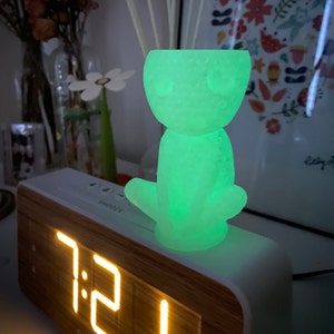 May include: A green glow-in-the-dark figurine of a creature with large eyes sitting on top of a white digital clock that displays the time 7:21.