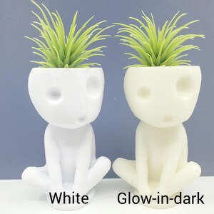 Puede incluir: Dos peque&ntilde;as macetas blancas con forma de figura sentada con una planta verde en cada una. La maceta de la izquierda es blanca y la maceta de la derecha brilla en la oscuridad.