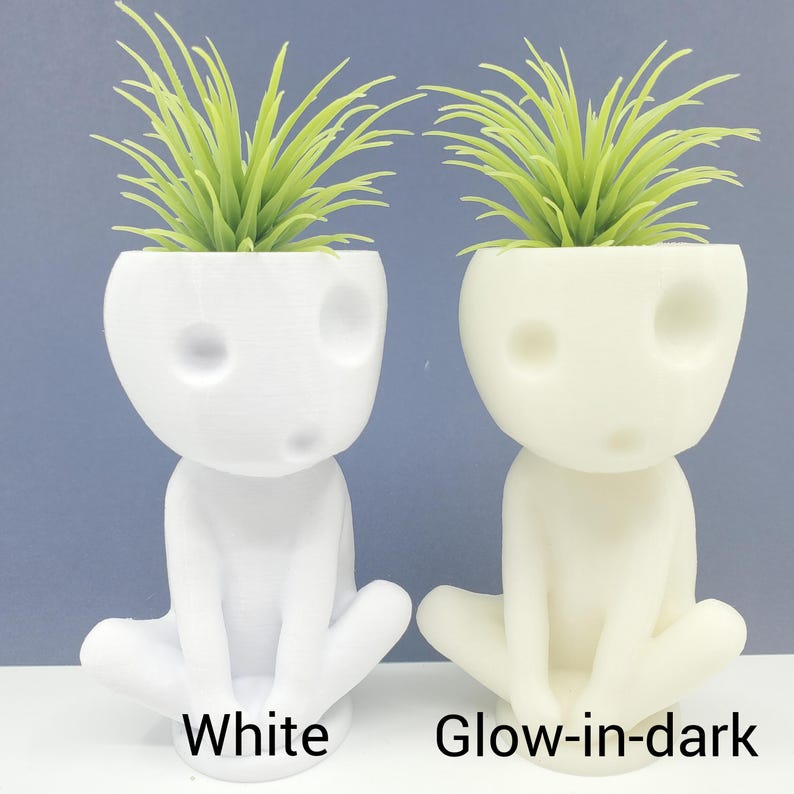 Puede incluir: Dos peque&ntilde;as macetas blancas con forma de figura sentada con una planta verde en cada una. La maceta de la izquierda es blanca y la maceta de la derecha brilla en la oscuridad.