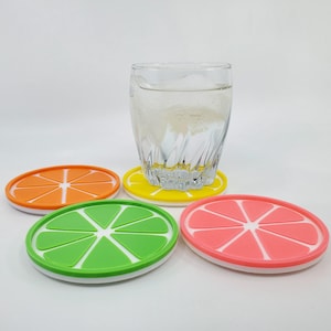 May include: Four silicone coasters shaped like citrus fruit slices. The coasters are orange, yellow, green, and pink. A glass of water sits on top of the yellow coaster.