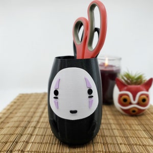 May include: Black pen holder with a white face design and lavender accents. A pair of red and gray scissors is inside the holder. A candle and a small fox-shaped planter are in the background.