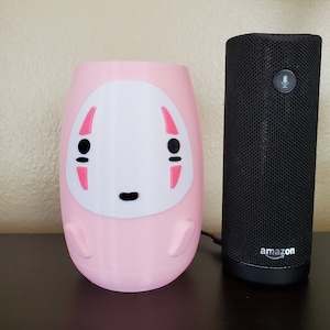 May include: A pink ceramic planter shaped like the No Face character from the animated film Spirited Away. The planter is next to a black Amazon Echo Dot smart speaker.