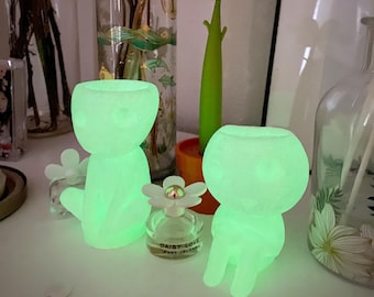 May include: Two glowing green ceramic figurines shaped like people, sitting on a white surface. The figurines are illuminated from within, creating a soft, ethereal glow. A small bottle of perfume with a white daisy on top is in front of the figurines.