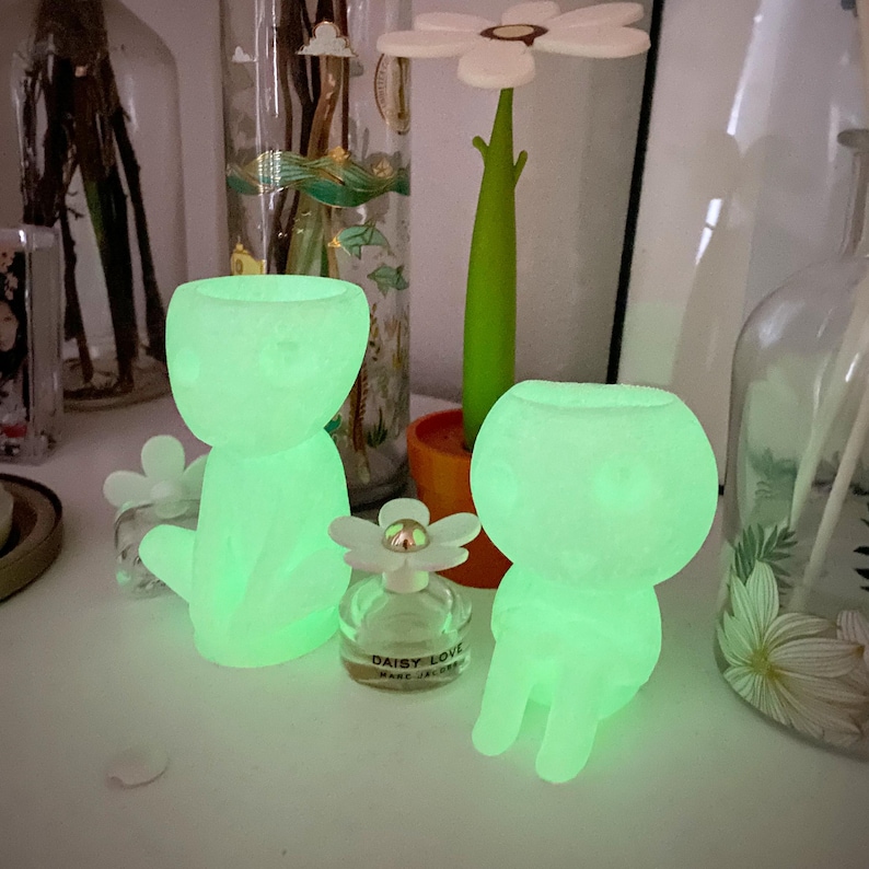 Puede incluir: Dos figuritas de cer&aacute;mica verde que brillan en la oscuridad, con forma de personas, sentadas sobre una superficie blanca. Las figuritas est&aacute;n iluminadas desde el interior, creando un brillo suave y et&eacute;reo. Un peque&ntilde;o frasco de perfume con una margarita blanca en la parte superior se encuentra delante de las figuritas.