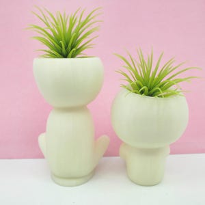 Puede incluir: Dos macetas de cer&aacute;mica blanca con forma de figuras humanas estilizadas, cada una con una planta de aire verde en su interior.