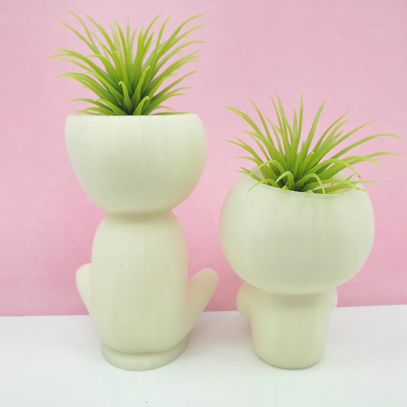 Puede incluir: Dos macetas de cer&aacute;mica blanca con forma de figuras humanas estilizadas, cada una con una planta de aire verde en su interior.