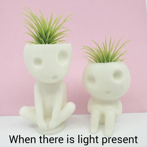Puede incluir: Dos macetas de cer&aacute;mica blanca con forma de figuras humanas con plantas verdes que crecen de sus cabezas. Las macetas est&aacute;n sobre una superficie rosa.  "When there is light present" est&aacute; escrito en negro en la parte inferior de la imagen.