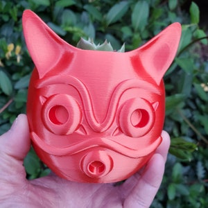 May include: A coral-colored, 3D-printed planter shaped like a stylized fox head. The planter has large, round eye openings, pointed ears, and a small nose opening. A small succulent plant is visible inside the planter.